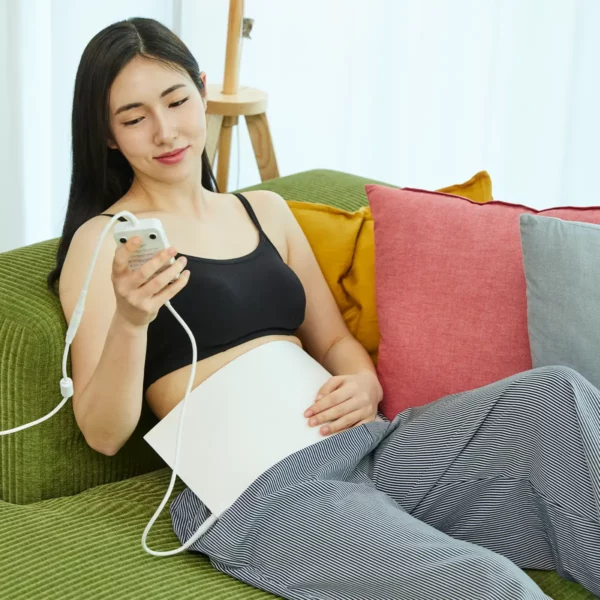 abdomen-irradiator-using-controller Woman sitting with the Abdomen Irradiator mat on her stomach, adjusting settings on the controller for red light therapy.